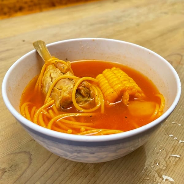 Cuban chicken soup with noodles, corn on the cob, and a chicken drumstick in a white bowl.