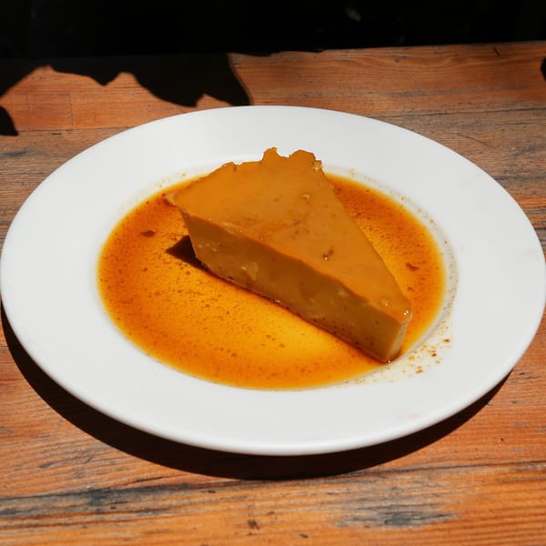 A slice of golden flan with caramel sauce on a white plate, on a wooden table.